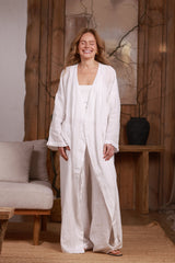 Front view of a long white linen kimono worn open over a white linen set. This elegant and breathable kimono is perfect for summer beach days, resort lounging, and casual outings in Dubai, UAE, and GCC. The minimalist wooden decor enhances the natural, sustainable aesthetic.