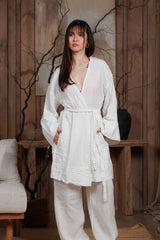 Front view of a white short muslin robe layered over white linen trousers. This lightweight and breathable combination is designed for the hot climates of Dubai, UAE, and GCC. Ideal for beachwear, resort styling, and summer city looks. Styled in a minimalist, natural-themed interior, reflecting sustainable Middle Eastern elegance.