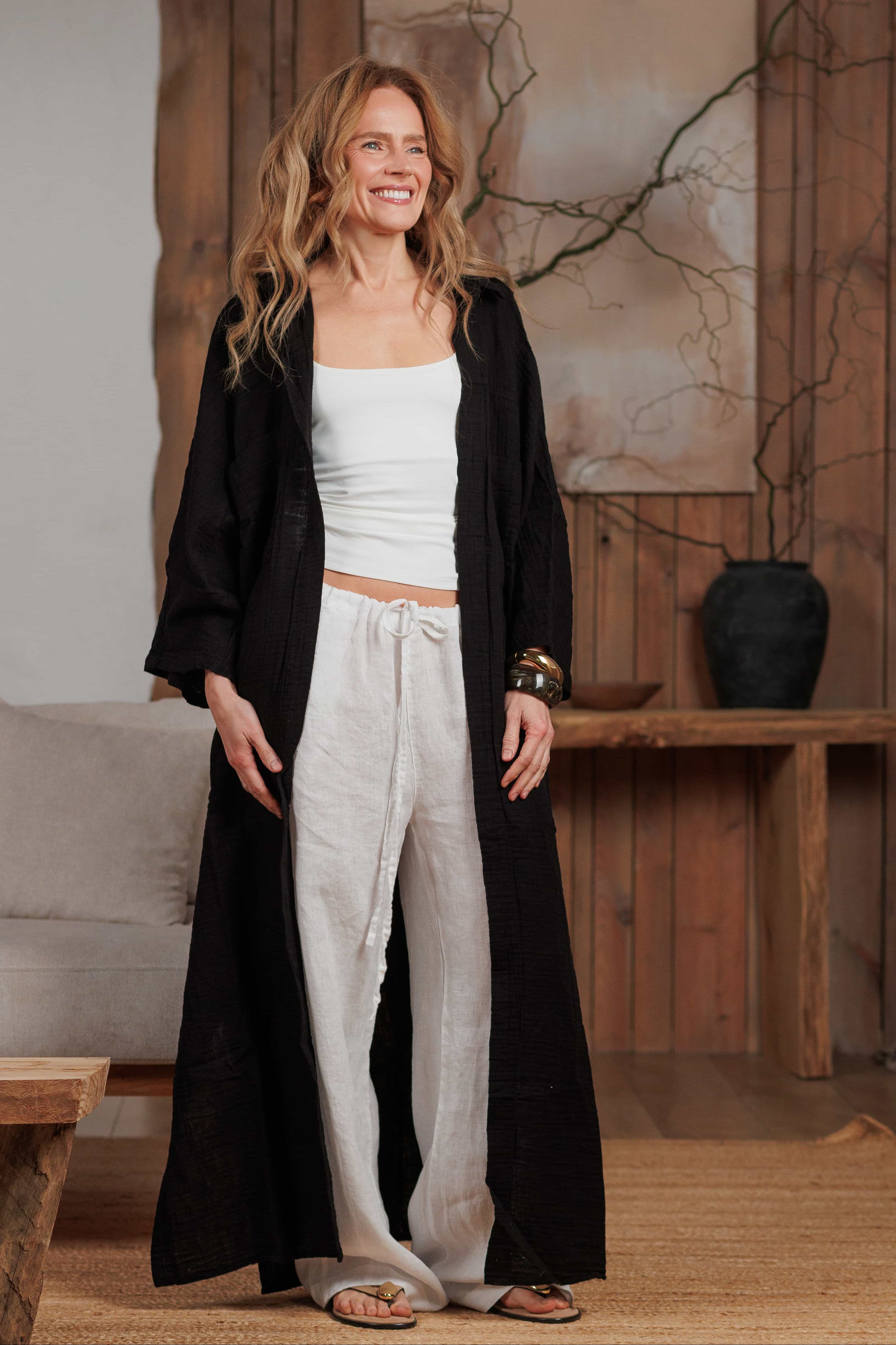 Woman wearing a long black muslin shirt over white linen trousers, standing in a minimalist boho-style living room. The lightweight black muslin shirt flows elegantly, perfect for beach or casual city wear in warm climates. Ideal for GCC markets looking for breathable, sustainable fashion.