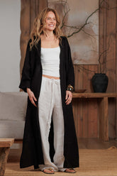 Woman wearing a long black muslin shirt over white linen trousers, standing in a minimalist boho-style living room. The lightweight black muslin shirt flows elegantly, perfect for beach or casual city wear in warm climates. Ideal for GCC markets looking for breathable, sustainable fashion.