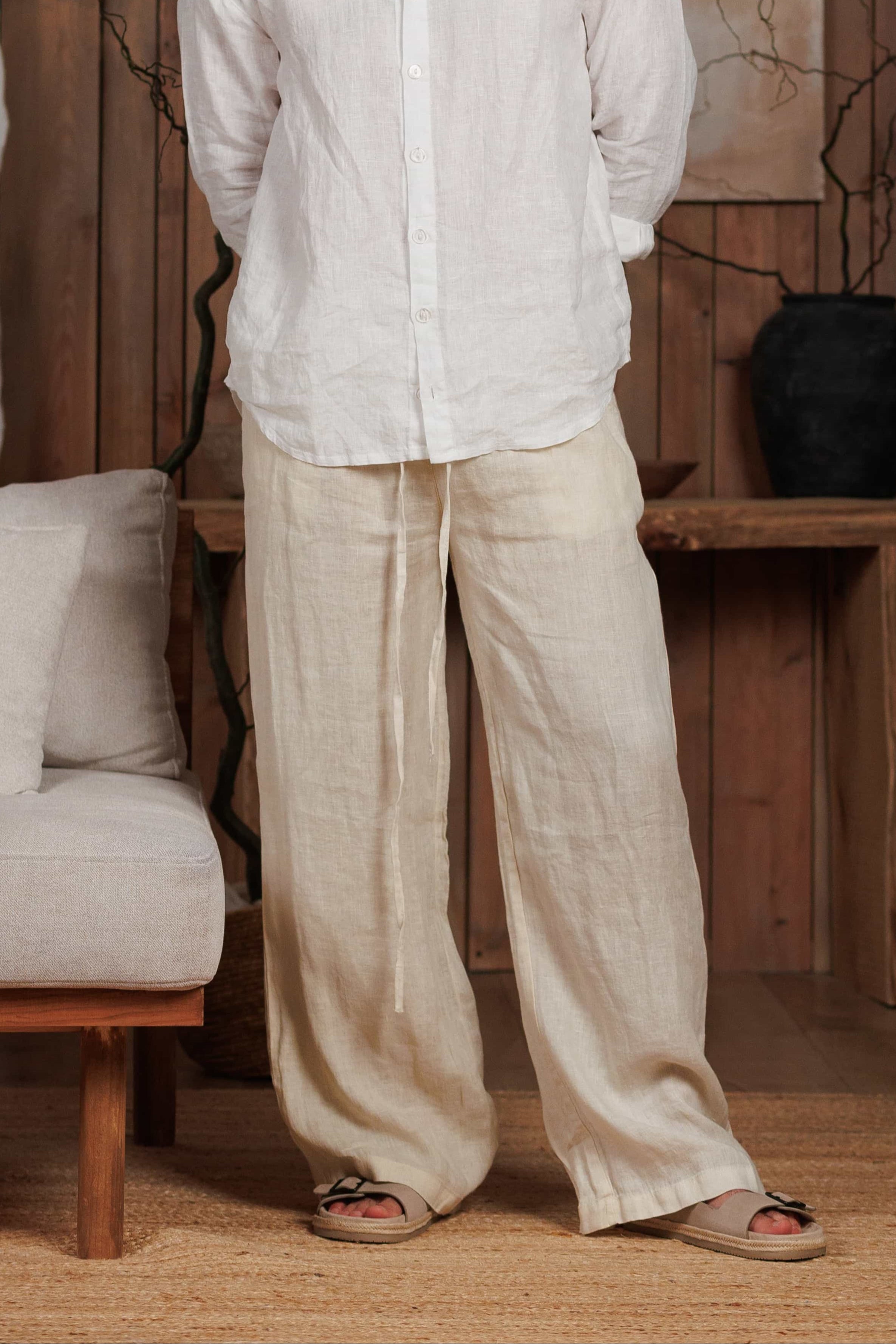 ront view of a men's white linen shirt paired with beige linen wide-leg trousers, designed for the hot climates of Dubai, UAE, and GCC. The breathable and lightweight linen fabric ensures comfort and elegance for beach, city, and casual wear. Styled in a minimalist, nature-inspired setting with wooden accents, reflecting sustainable Middle Eastern fashion.