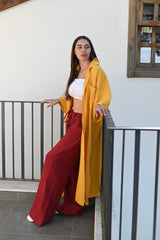 Woman wearing a long amber muslin shirt styled with burgundy linen trousers and a white muslin crop top, standing gracefully on a balcony with a minimalist white backdrop. Perfect for summer outings and effortless elegance in the GCC region.