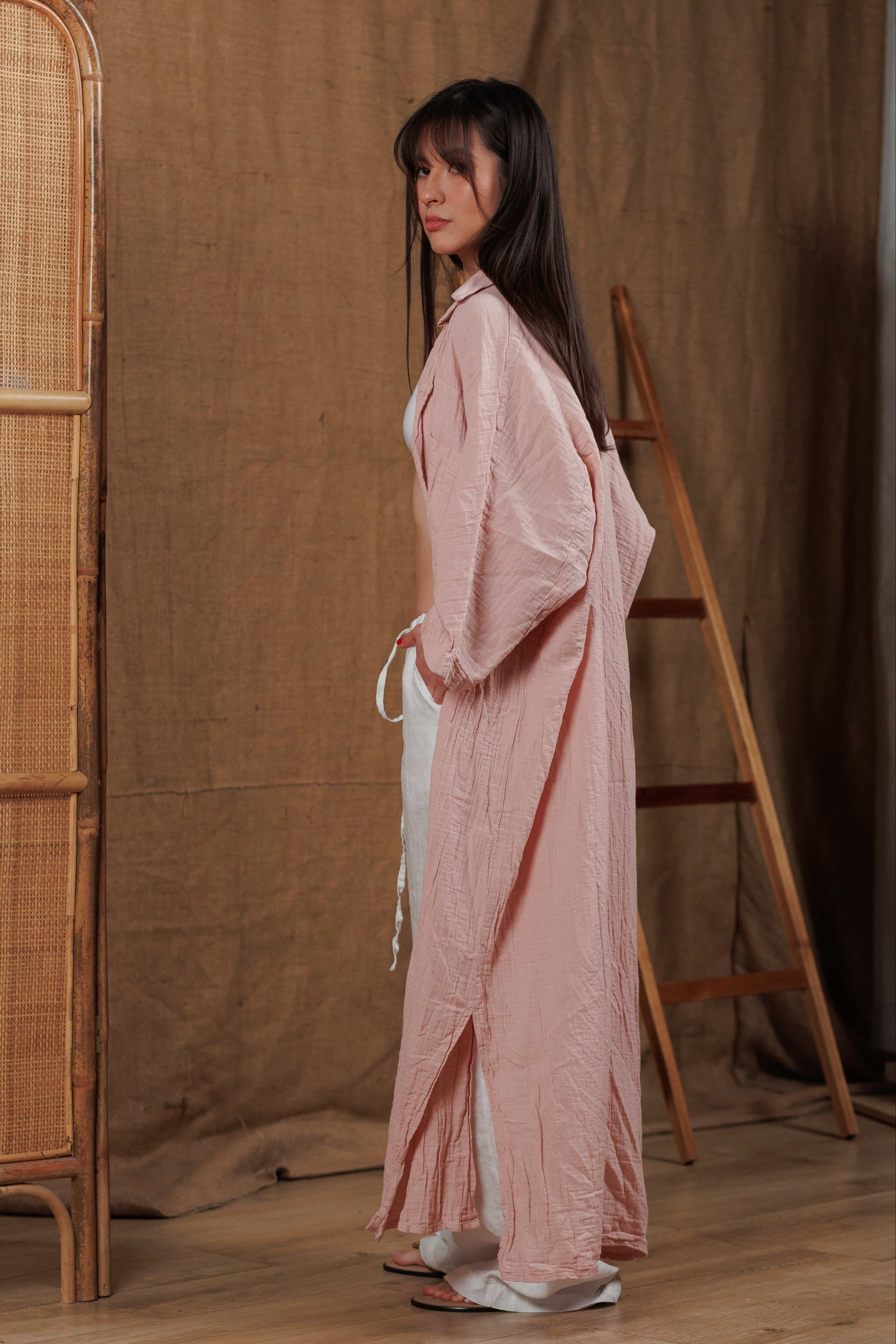 Side view of a long muslin women's shirt in a delicate tea rose shade, paired with white linen trousers. The lightweight fabric flows effortlessly, making it perfect for summer outings and beachwear in hot climates like the UAE