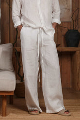 White linen men's trousers with a relaxed fit and adjustable drawstring waist, ideal for hot climates like Dubai, the UAE, and GCC countries. Styled in a minimalist, natural-themed setting with wooden accents, these breathable and sustainable linen pants are perfect for beach, city, and home wear. Discover lightweight trousers designed for Dubai summer fashion and Middle Eastern elegance.
