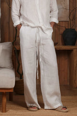 White linen men's trousers with a relaxed fit and adjustable drawstring waist, ideal for hot climates like Dubai, the UAE, and GCC countries. Styled in a minimalist, natural-themed setting with wooden accents, these breathable and sustainable linen pants are perfect for beach, city, and home wear. Discover lightweight trousers designed for Dubai summer fashion and Middle Eastern elegance.
