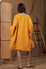 Back view of a lightweight yellow amber muslin robe, designed for the warm climates of Dubai and GCC. Its relaxed fit and breathable fabric make it ideal for beach cover-ups and resort wear. Photographed in a natural-themed interior with rattan accents, reflecting Middle Eastern elegance and sustainable fashion.