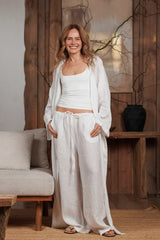Front view of a long white linen kimono styled over white linen trousers and a white tank top. This breathable and lightweight ensemble is ideal for summer days in Dubai, UAE, and across the GCC. Perfect for beach cover-ups, resort wear, and casual city looks. The minimalist wooden interior reflects Middle Eastern elegance and sustainable fashion.