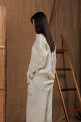 Side view of a woman wearing a beige linen shirt and wide-leg linen pants, styled in a rustic setting with natural wooden and woven decor. The shirt's relaxed fit and breathable linen fabric make it perfect for the warm climate of the UAE and GCC region. Ideal for casual and resort wear.