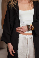 Close-up of a long black muslin shirt paired with white linen trousers. The textured fabric and relaxed fit make it an essential layering piece for warm weather, appealing to the GCC market with its minimalist design and breathable materials.