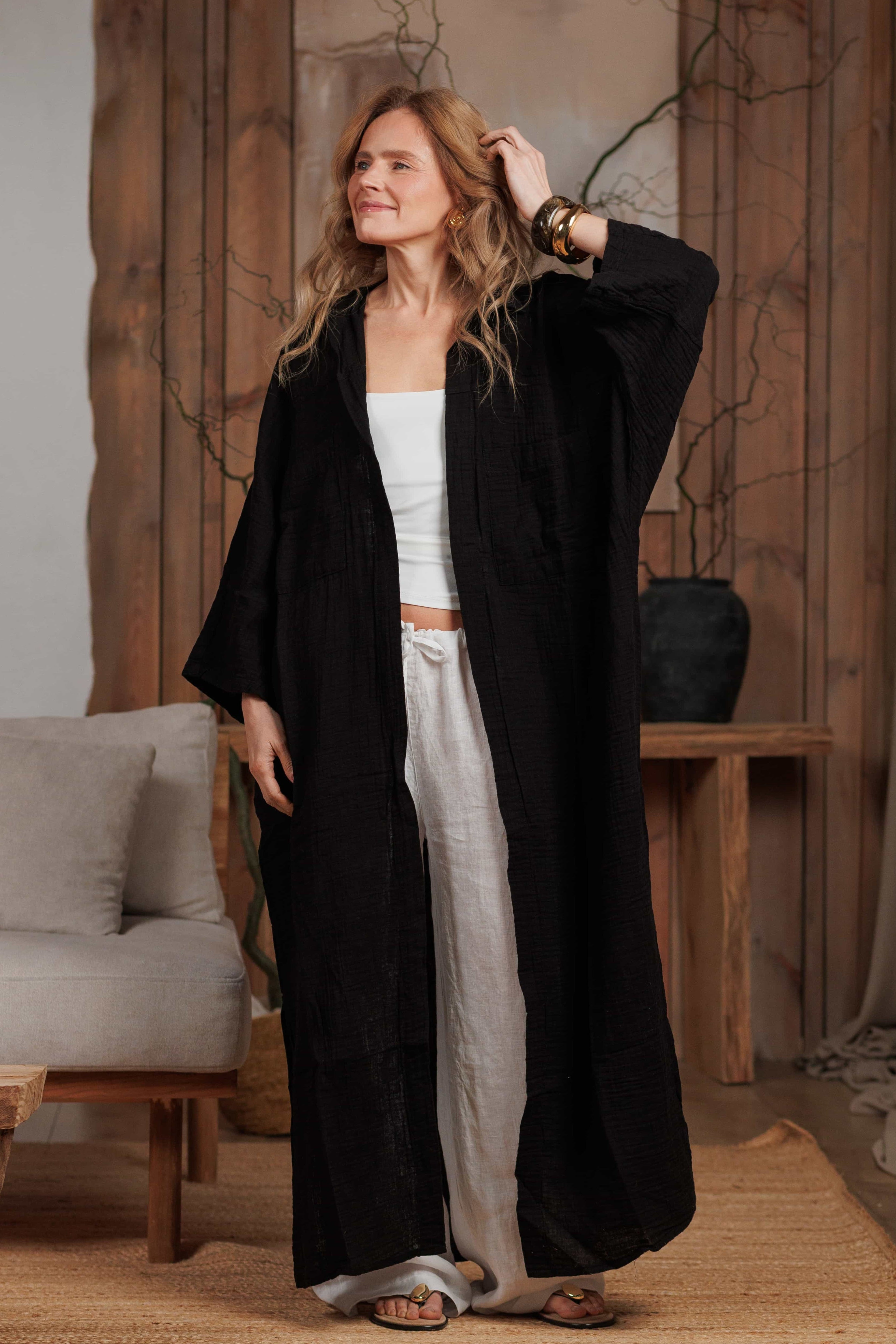 Woman posing in a long black muslin shirt and white linen trousers, accessorized with bold bracelets. The outfit reflects effortless elegance, designed for beach, home, and city wear in hot climates. Crafted from 100% natural fabrics, ideal for sustainable fashion enthusiasts in the Middle East.