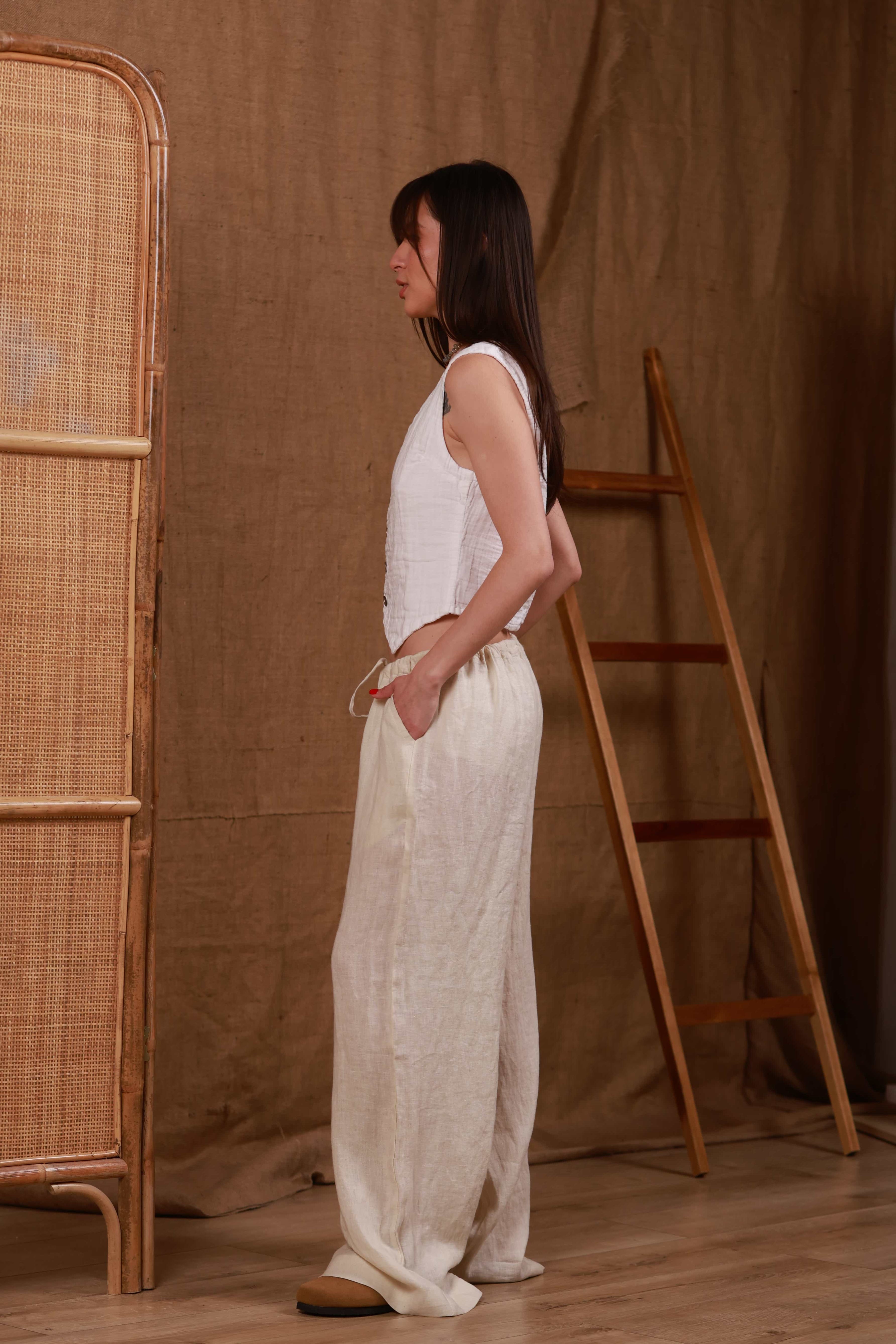 Side view of beige women's linen trousers paired with a white muslin top. The relaxed fit and natural fabric emphasize comfort and breathability, perfect for summer days in the UAE. The look is completed with minimalist accessories, ideal for beach or resort wear.