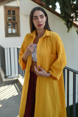 Woman elegantly adjusting her long amber muslin shirt over a white muslin crop top and burgundy linen trousers, styled with minimalistic jewelry. Ideal for beachwear, resort fashion, and summer city walks in the Middle East.