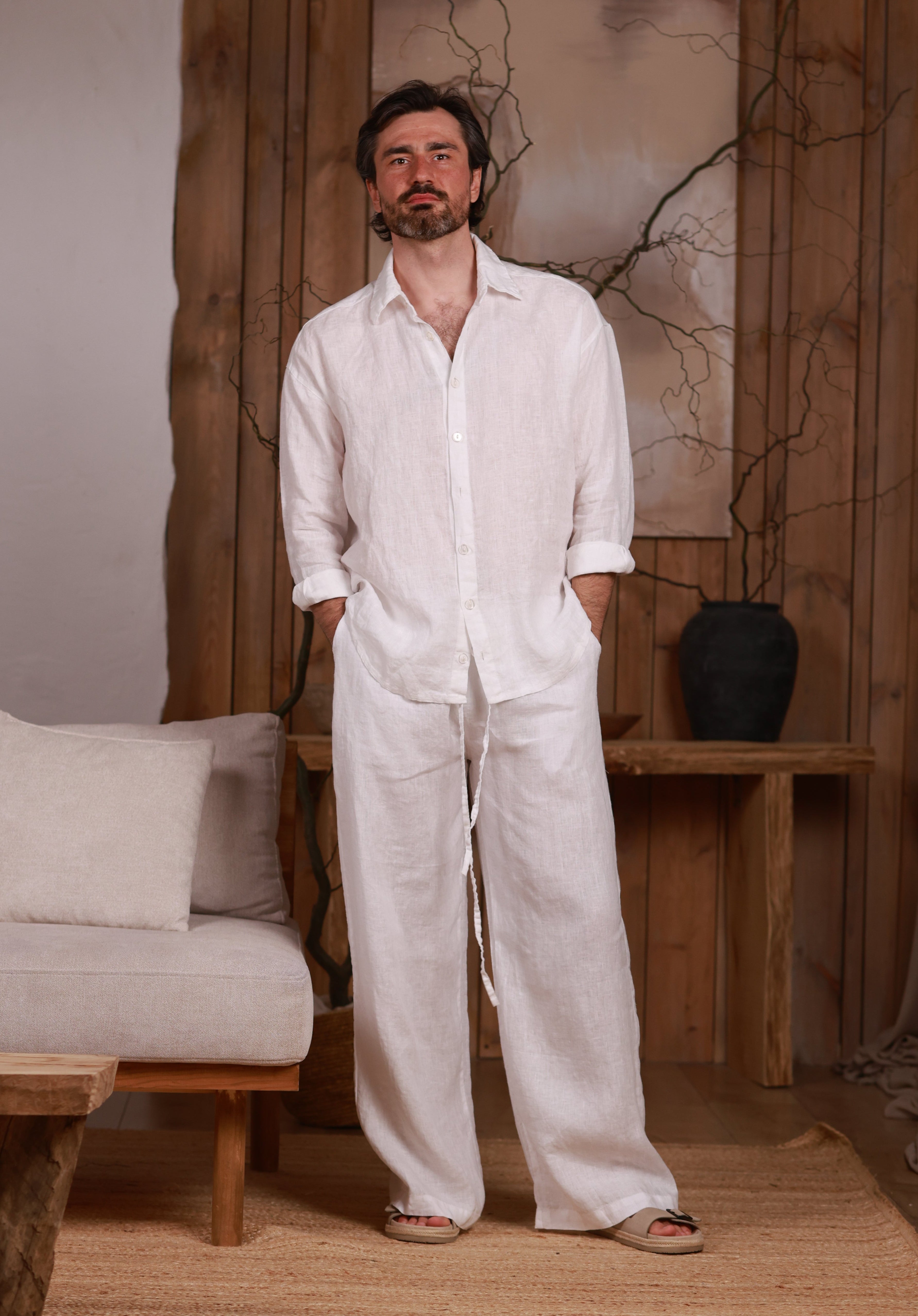 White linen men's shirt paired with wide-leg linen trousers. Linen white set. The model is standing in a minimalist, natural-themed room with wooden accents and neutral-toned furniture, reflecting a relaxed and sophisticated look.