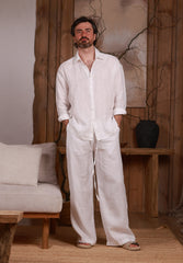 White linen men's shirt paired with wide-leg linen trousers. Linen white set. The model is standing in a minimalist, natural-themed room with wooden accents and neutral-toned furniture, reflecting a relaxed and sophisticated look.
